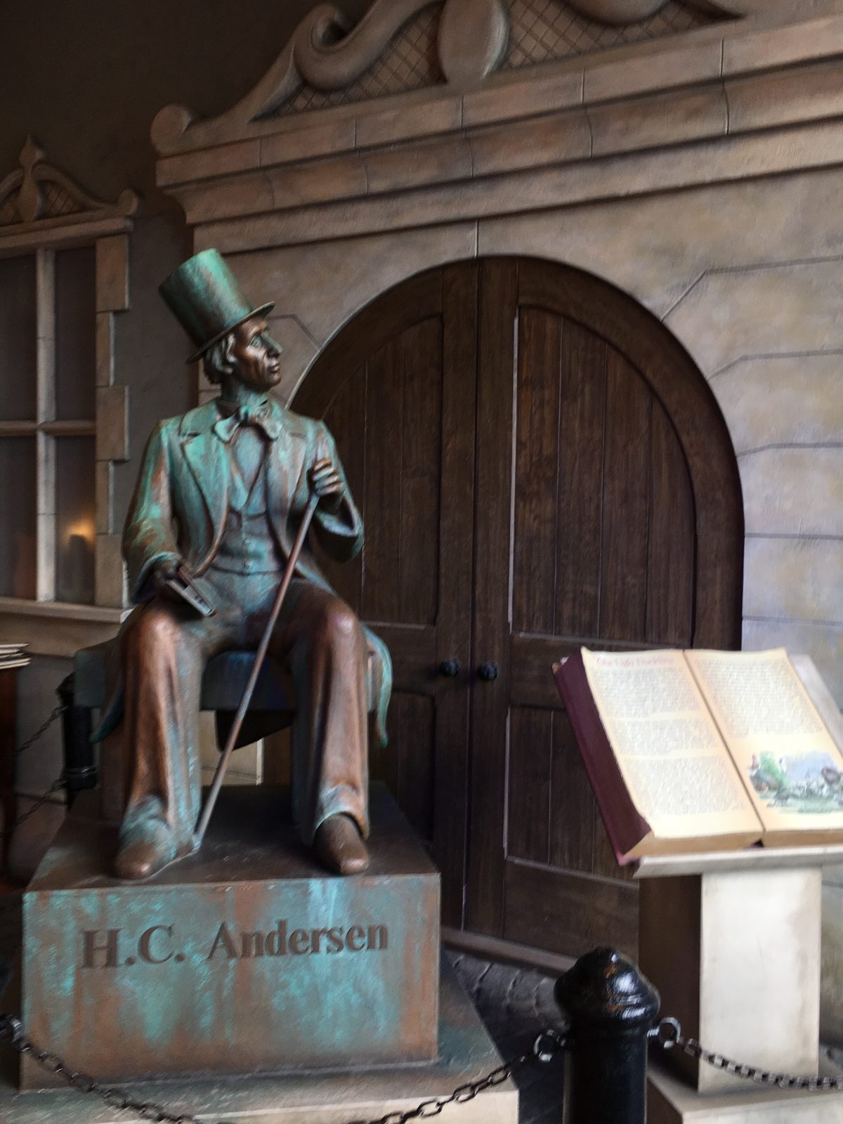A bronze statue of Hans Christian Andersen wearing a top hat and holding a cane, seated before an arched wooden door with an open storybook on a stand beside it.
