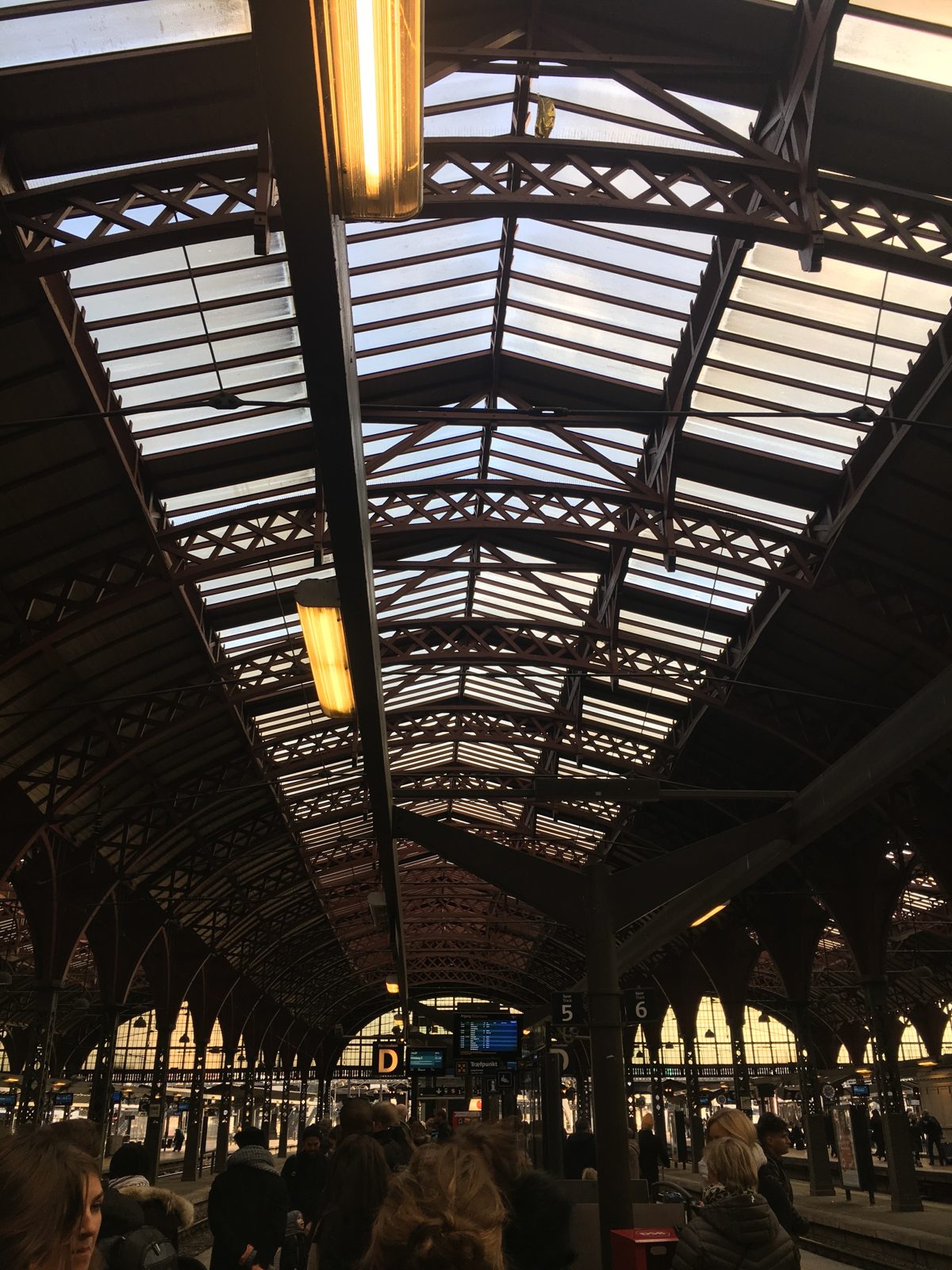 The intricate wooden roof trusses and glass skylights of Copenhagen Central Station, with platform signs and travelers below.