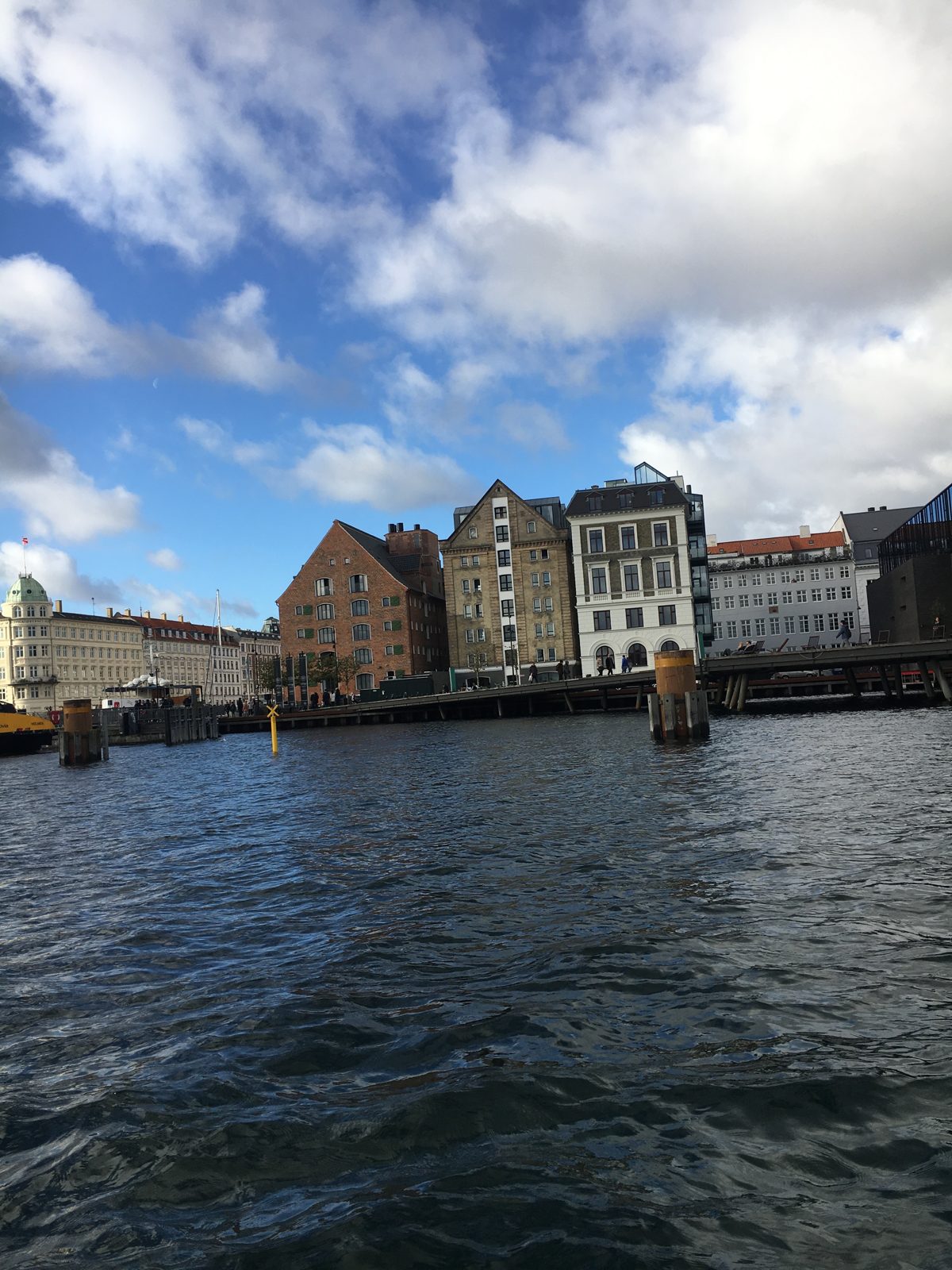 Brick and stucco waterfront warehouses and townhouses along a Copenhagen canal, with rippling water in the foreground and bright clouds above.
