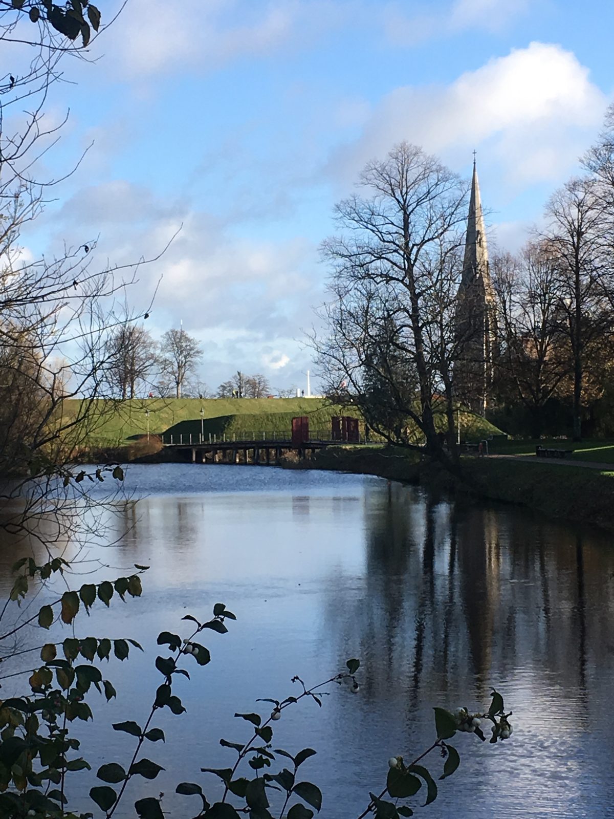 The slender spire of St. Alban's Church reflected in the calm moat water at Kastellet, framed by autumn trees and a grassy rampart.