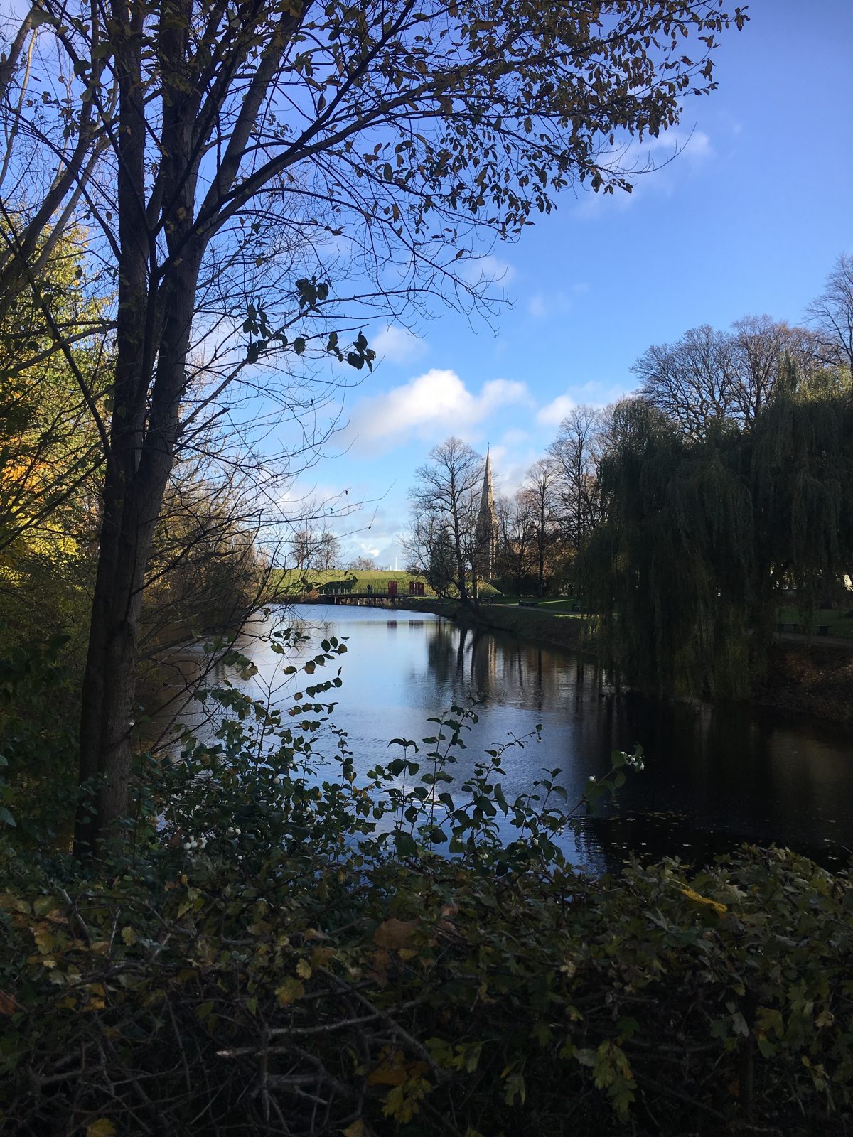 The spire of St. Alban's Church seen through autumn-leaved branches across the still water of the Kastellet moat.