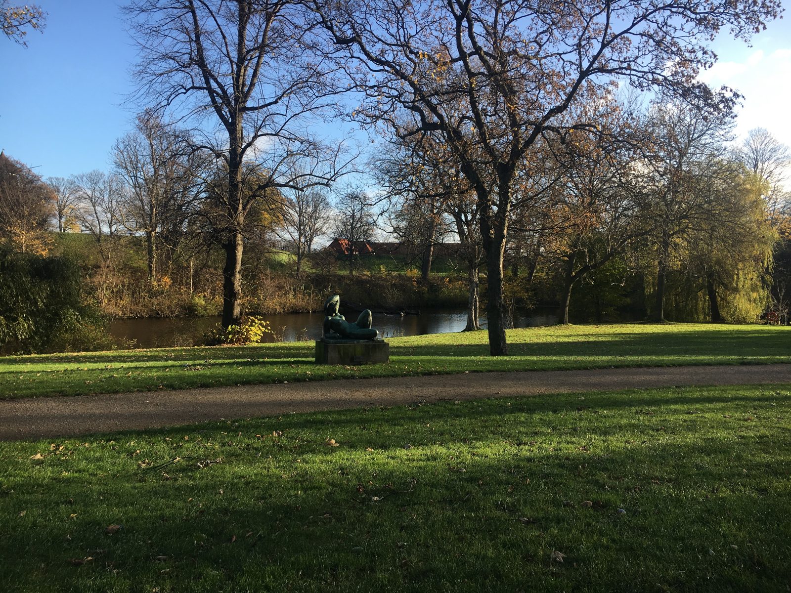 A reclining bronze sculpture on a stone plinth in a sunlit lawn at Kastellet park, beside a pond and bare autumn trees.