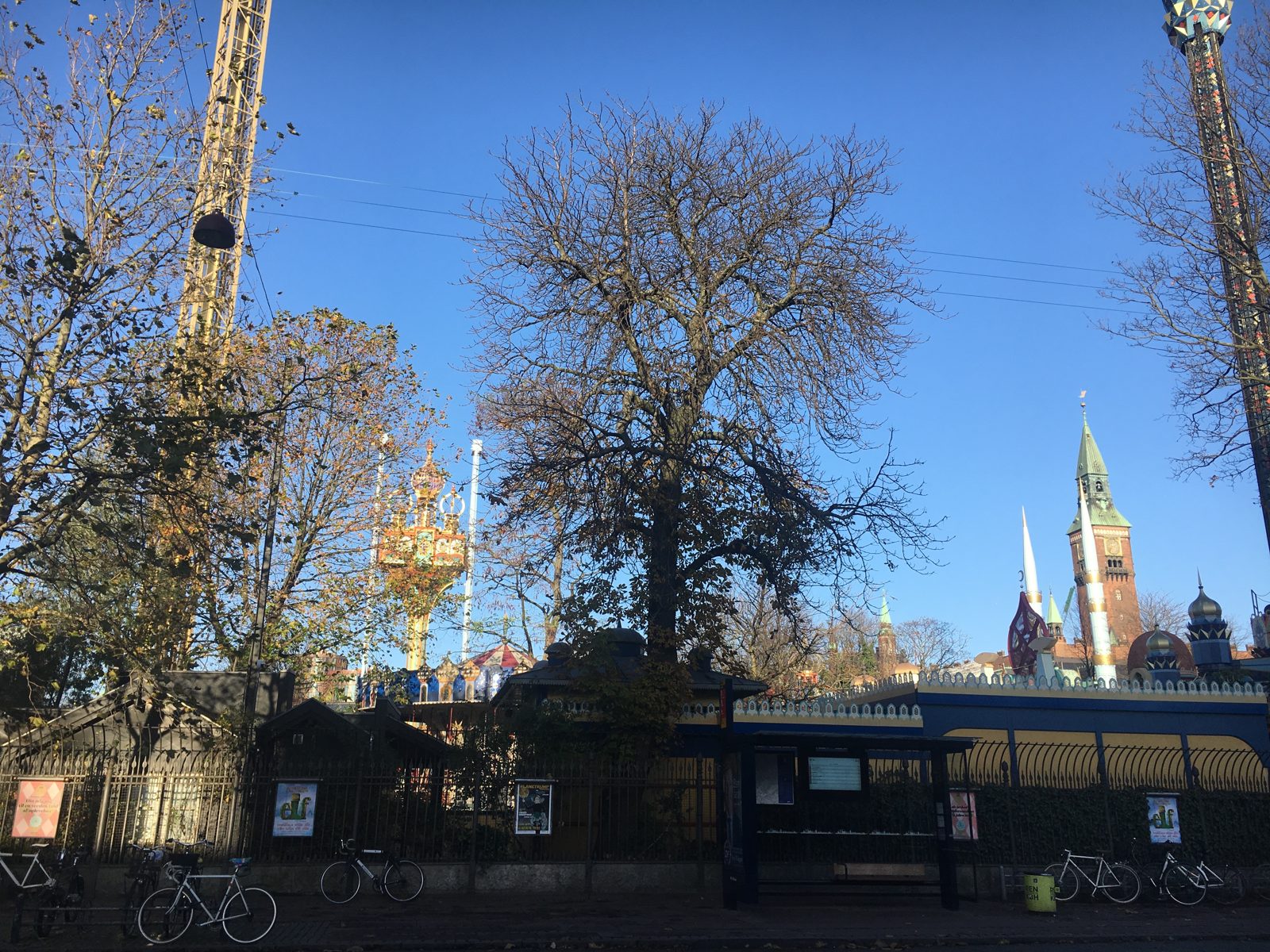 Colorful Tivoli Gardens ride towers and an ornate pavilion seen over a fence beside the green spire of Copenhagen City Hall under a clear blue sky.