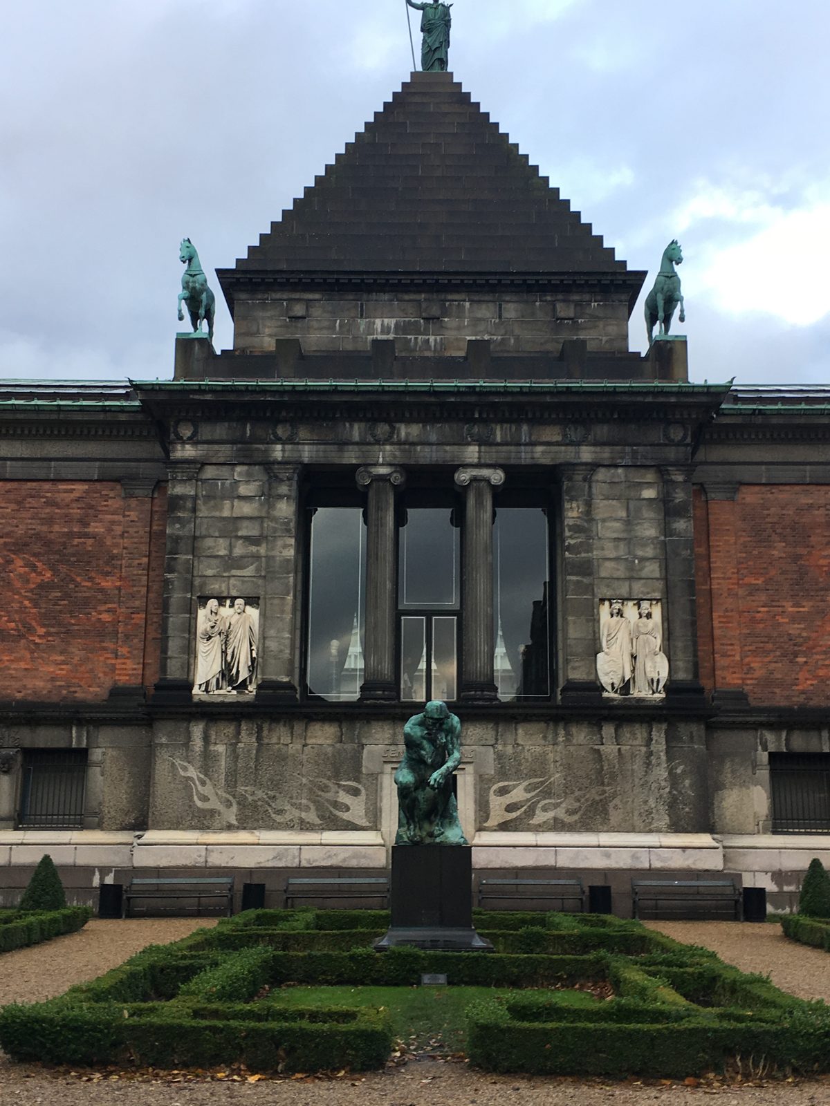 The pyramid-roofed facade of the Ny Carlsberg Glyptotek museum with bronze horse sculptures, a cast of Rodin's The Thinker, and a manicured boxwood garden in front.